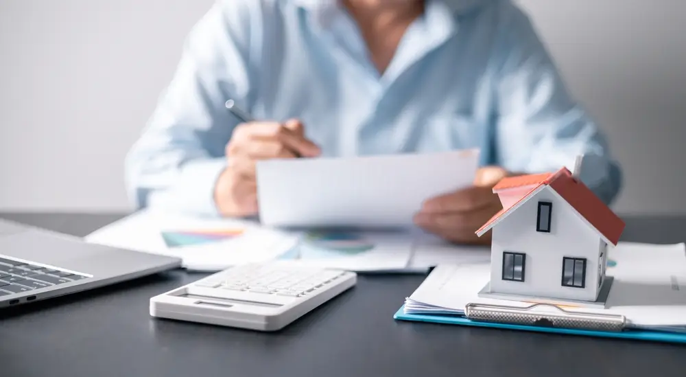 Man in a blue shirt reviews financial documents near a small house model, illustrating the Augusta Rule concept.