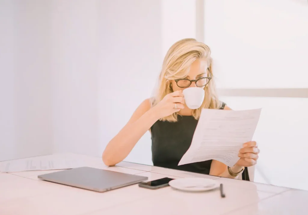 Blonde woman wearing glasses sips coffee and reviews tax extension documents for MyTax.Partners’